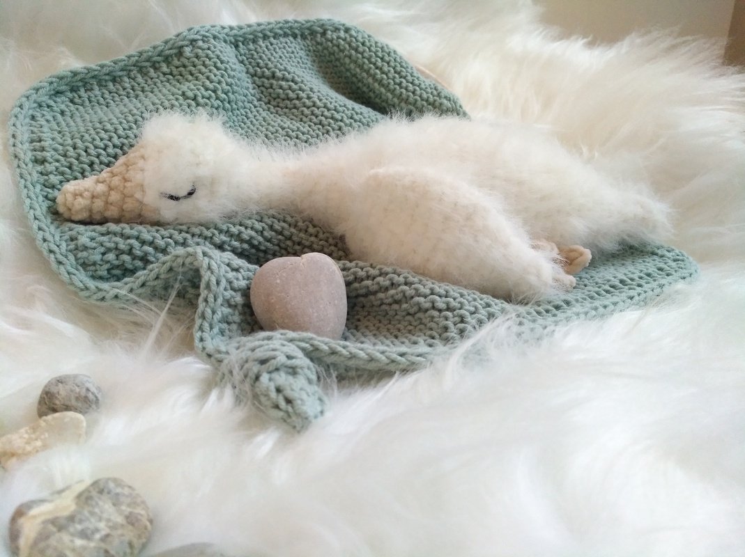 Gehäkelter weißer Schwan mit beigem Schnabel und geschlossenen Augen auf mintgrüner Strickdecke und weißem Plüsch.