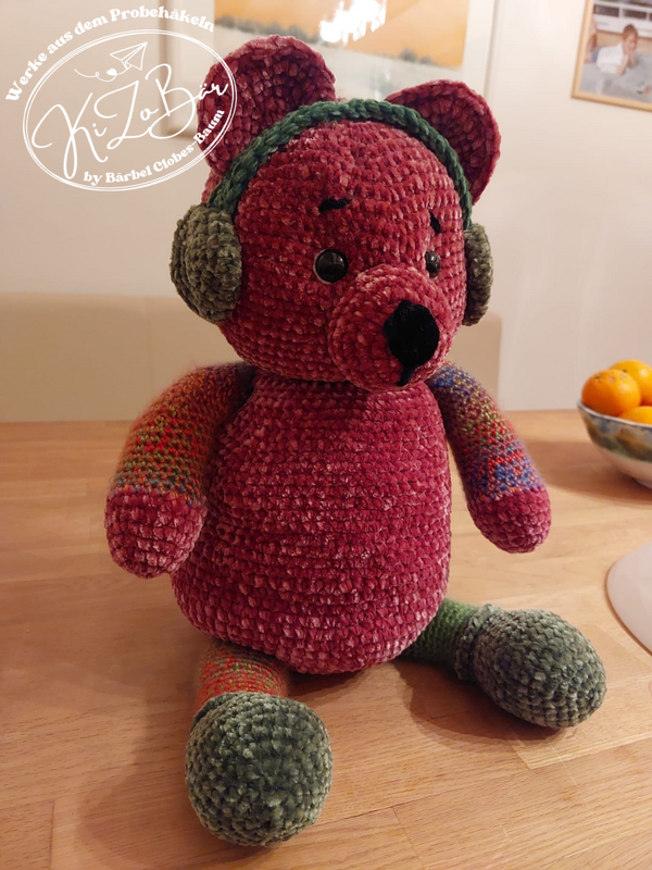 Dunkelroter Amigurumi-Teddybär aus Plüschgarn mit grünen Ohrenschützern, melierten Armen und grünen Füßen.
