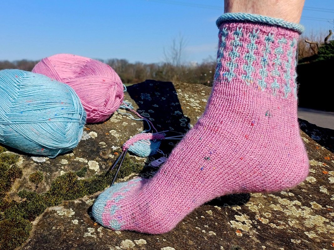 Fuß mit rosa handgestrickter Socke mit hellblauem Muster, Wollknäuel auf einem Stein