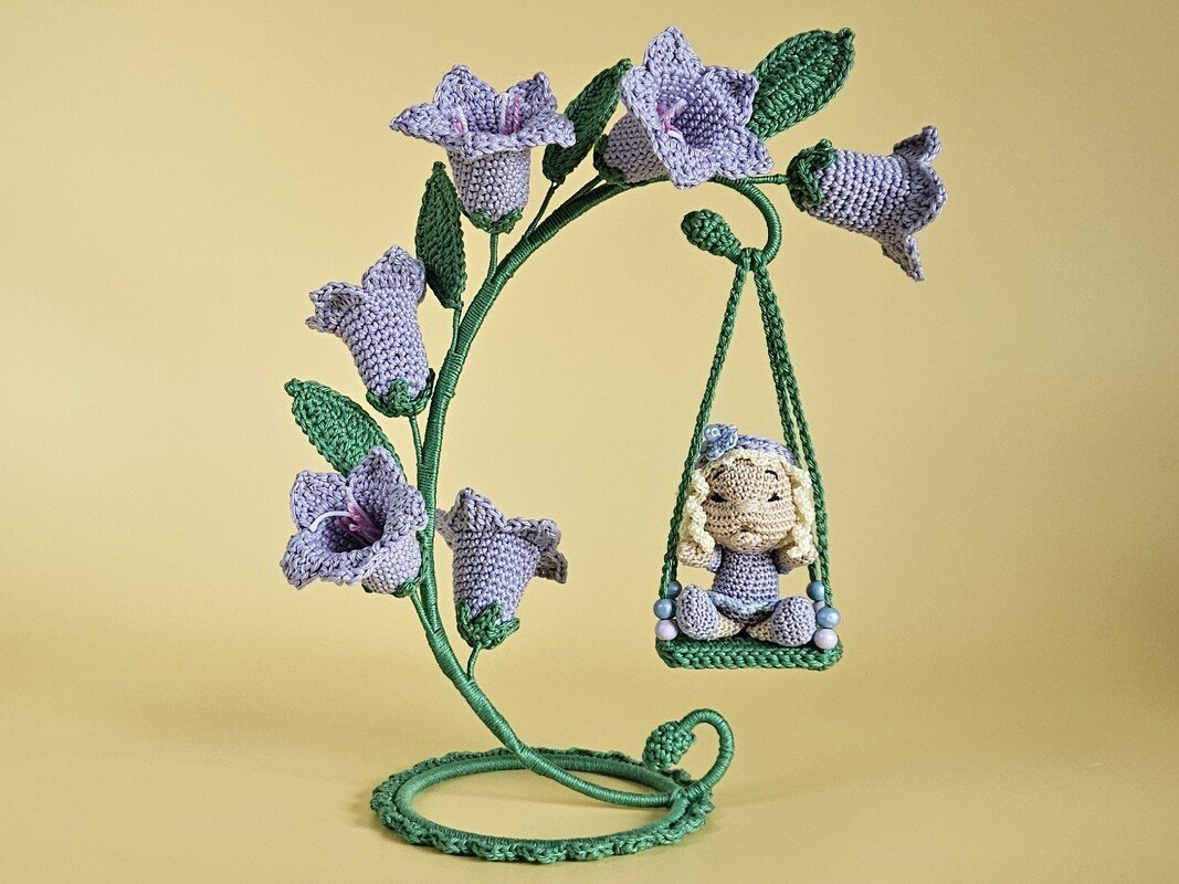 Gehäkelte Amigurumi-Puppe mit blonden Zöpfchen auf grüner Schaukel an einem Stiel mit violetten Glockenblüten