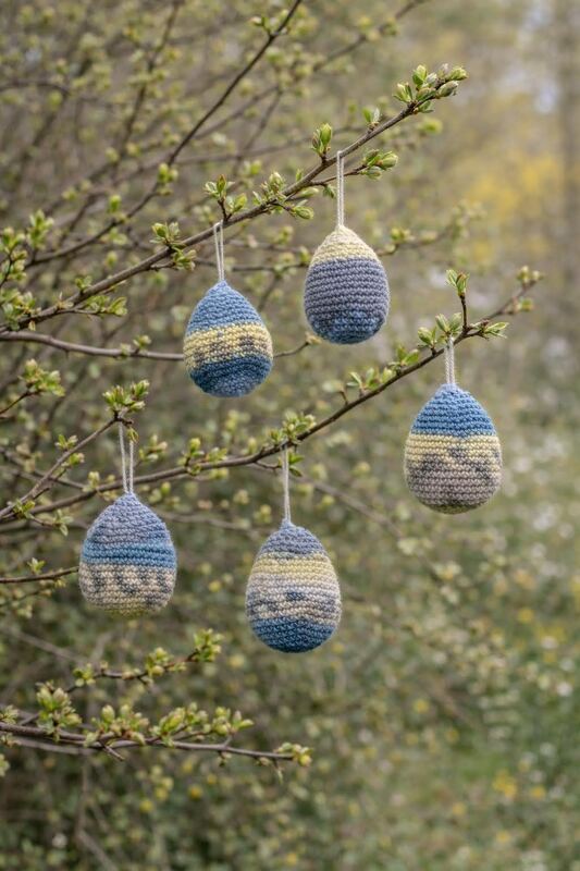 Fünf gehäkelte Ostereier in Blau und Creme an einem Zweig mit Knospen