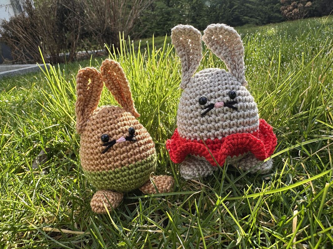 Zwei gehäkelte Osterhasen in Eiform im grünen Gras, einer braun-grün und einer beige mit roter Rüsche.