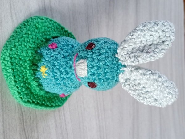 Türkiser Amigurumi-Hase mit weißen Ohren und farbigen Stickdetails auf grünem Kissen