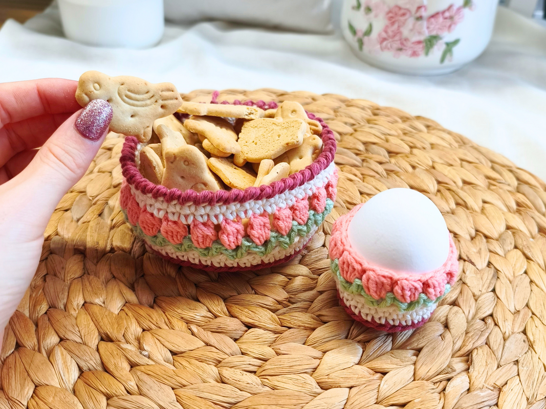 Gehäkeltes Körbchen mit Tulpenmuster, gefüllt mit Keksen in Tierform, daneben ein passender gehäkelter Eierbecher mit weißem Ei auf einem geflochtenen Platzset.