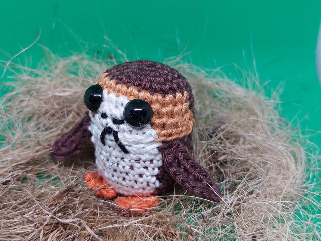Gehäkeltes Amigurumi-Vogelküken mit großen schwarzen Augen und orangefarbenen Füßen in einem Nest aus trockenem Gras vor grünem Hintergrund.