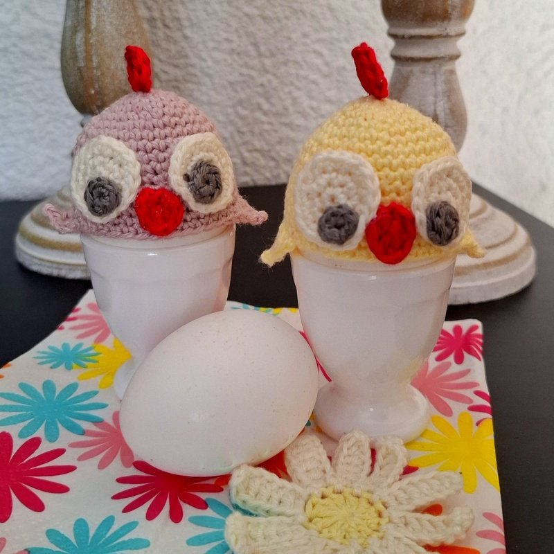 Gehäkelte Küken-Eierwärmer (pastellgelb und rosa) auf weißen Eierbechern mit Ei und Häkelblume