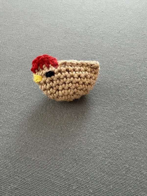 Gehäkeltes Küken-Amigurumi in Beige mit rotem Kamm und schwarzem Auge auf grauem Untergrund.