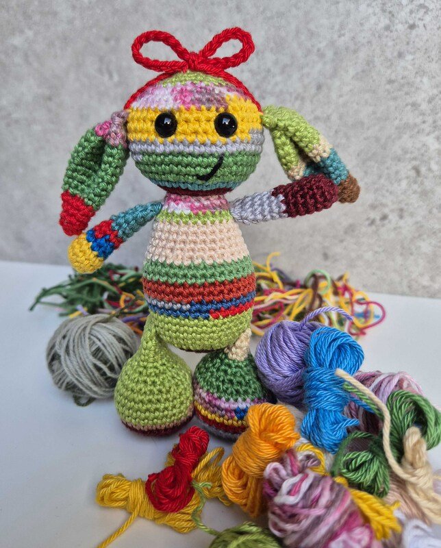 Bunte gehäkelte Amigurumi-Puppe mit roter Schleife, umgeben von Garnresten und Wollknäueln.