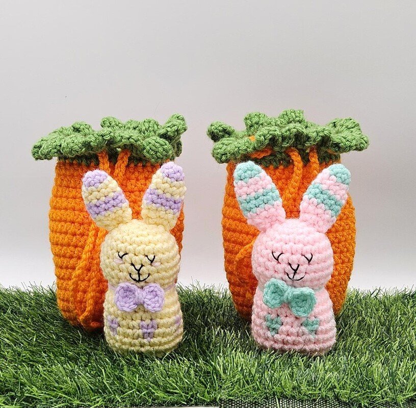 Two pastel crocheted amigurumi bunnies with orange carrot-shaped holders on grass