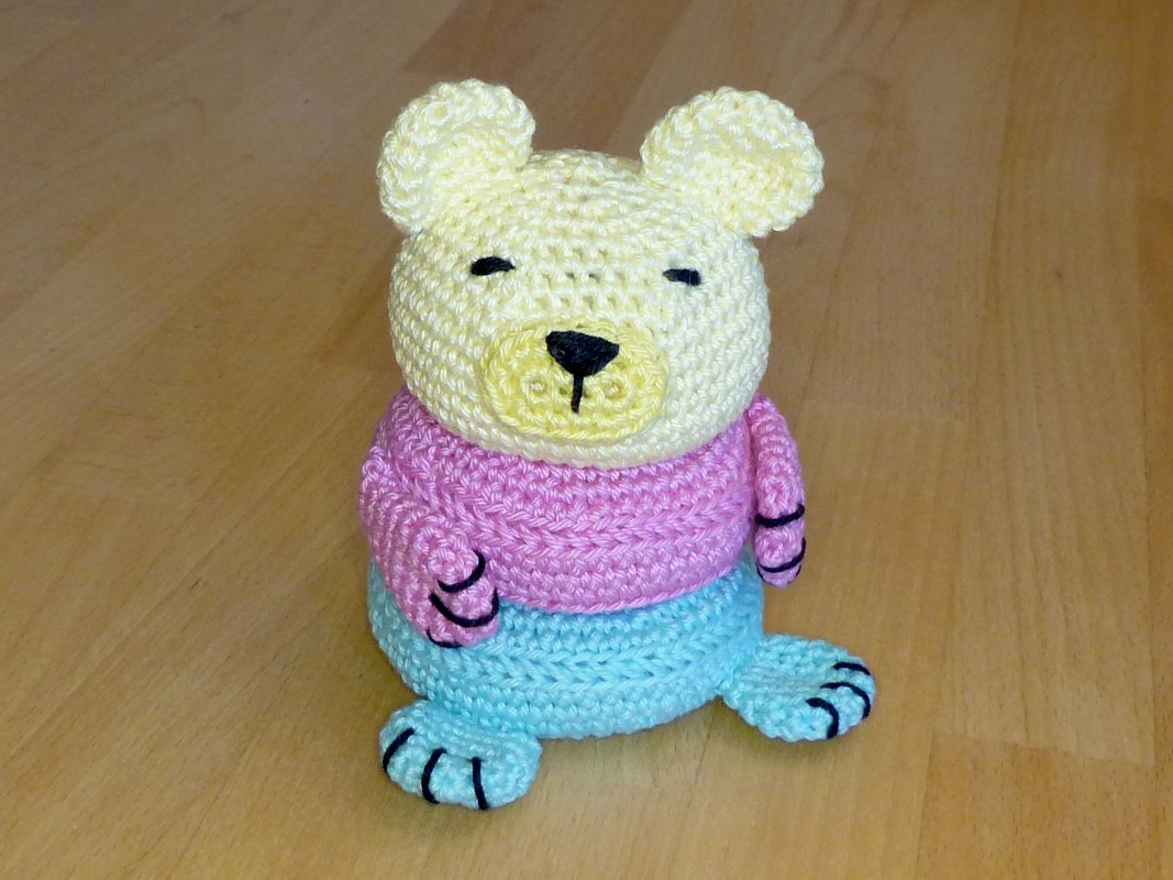 Sitzender gehäkelter Teddybär mit hellgelbem Kopf, rosa Oberteil und hellblauen Beinen auf Holzfußboden