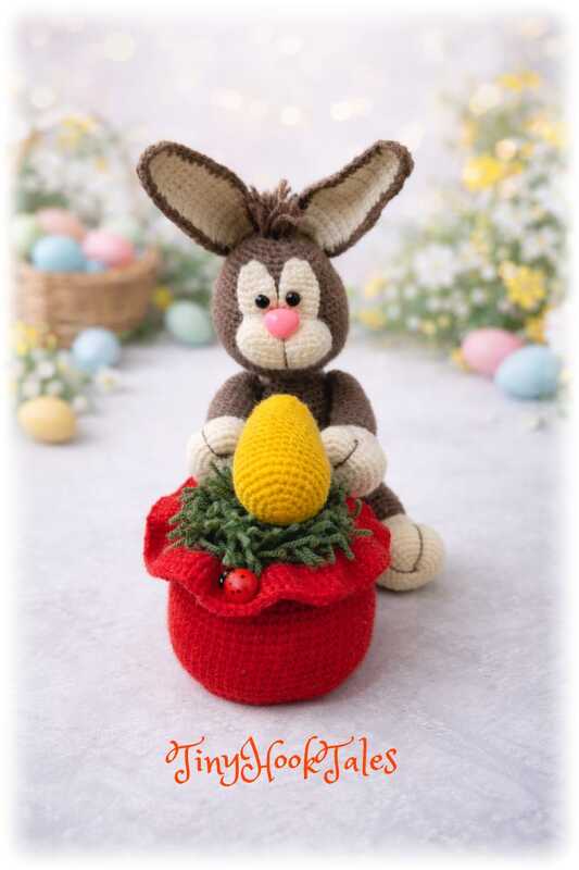Braun-beiger Amigurumi-Hase mit gelbem Ei in roter Hülle, unscharfer Hintergrund mit bunten Ostereiern