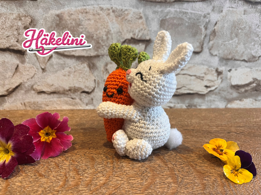 Gehäkelter weißer Amigurumi-Hase umarmt eine orange Möhre auf Holz, umgeben von bunten Frühlingsblüten