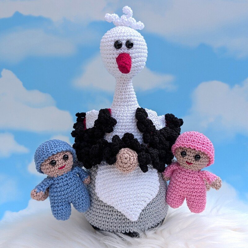 Gehäkelte Storchfigur hält zwei Amigurumi-Babys, eines blau, eines rosa, vor blau-weißem Wolkenhintergrund