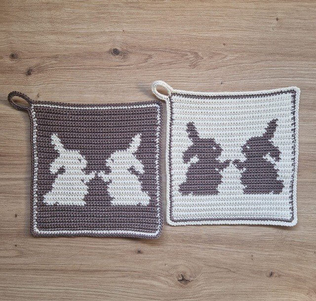 Zwei quadratische gehäkelte Topflappen mit farblich invertierten Hasenmotiven in Braun und Creme auf Holzuntergrund