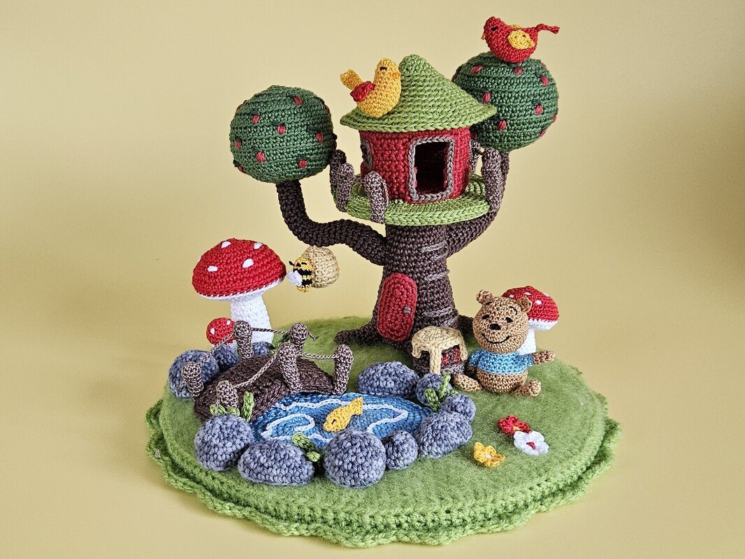 Amigurumi-Diorama mit Baumhaus im Baum, Teich mit Steinrand und Brücke, Fliegenpilzen, Vögeln, Biene und kleinem Bären auf grüner Basis.