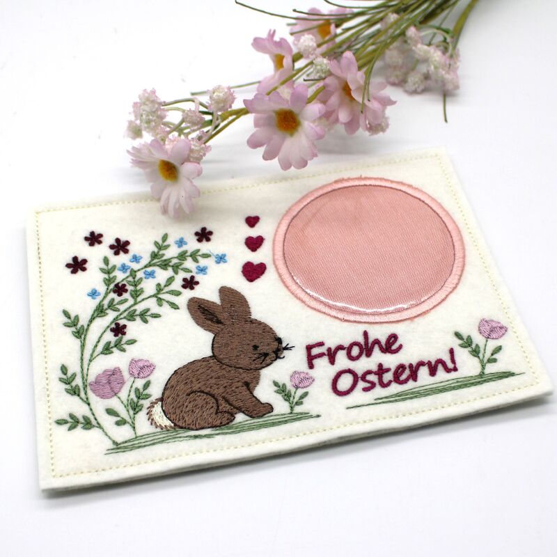 Bestickter Tassenteppich Ostern mit braunem Hasen, Blumen, Herzen und Schriftzug „Frohe Ostern!“ auf hellem Filz mit runder rosa Applikation