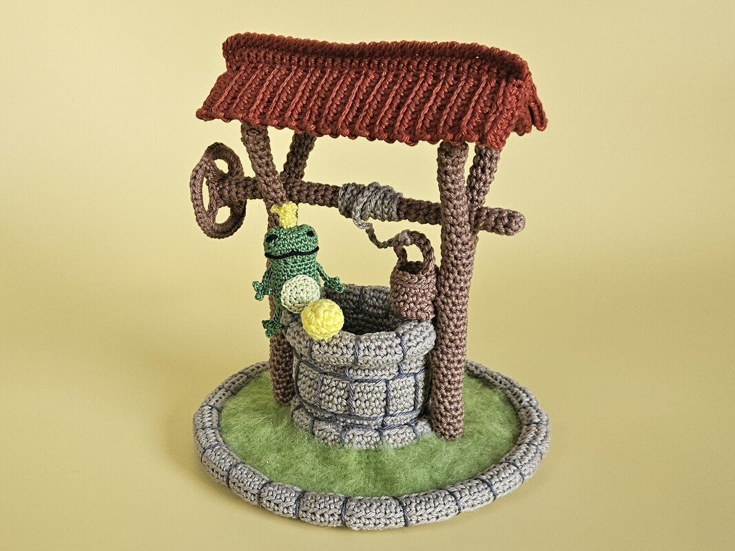 Gehäkelter Brunnen mit Dach, Kurbel und Frosch-Amigurumi auf rundem grünem Sockel