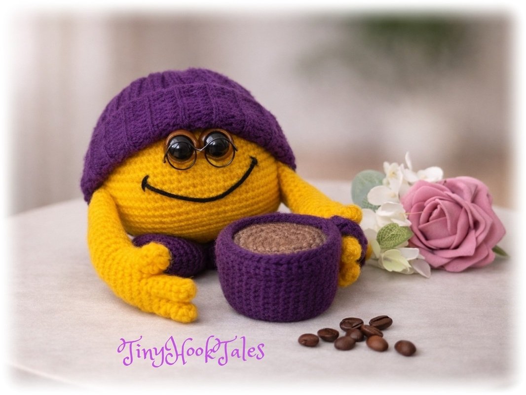 Gelbes gehäkeltes Smiley-Amigurumi mit violetter Mütze und Häkel-Tasse, Brille, Kaffeebohnen und rosafarbener Blüte