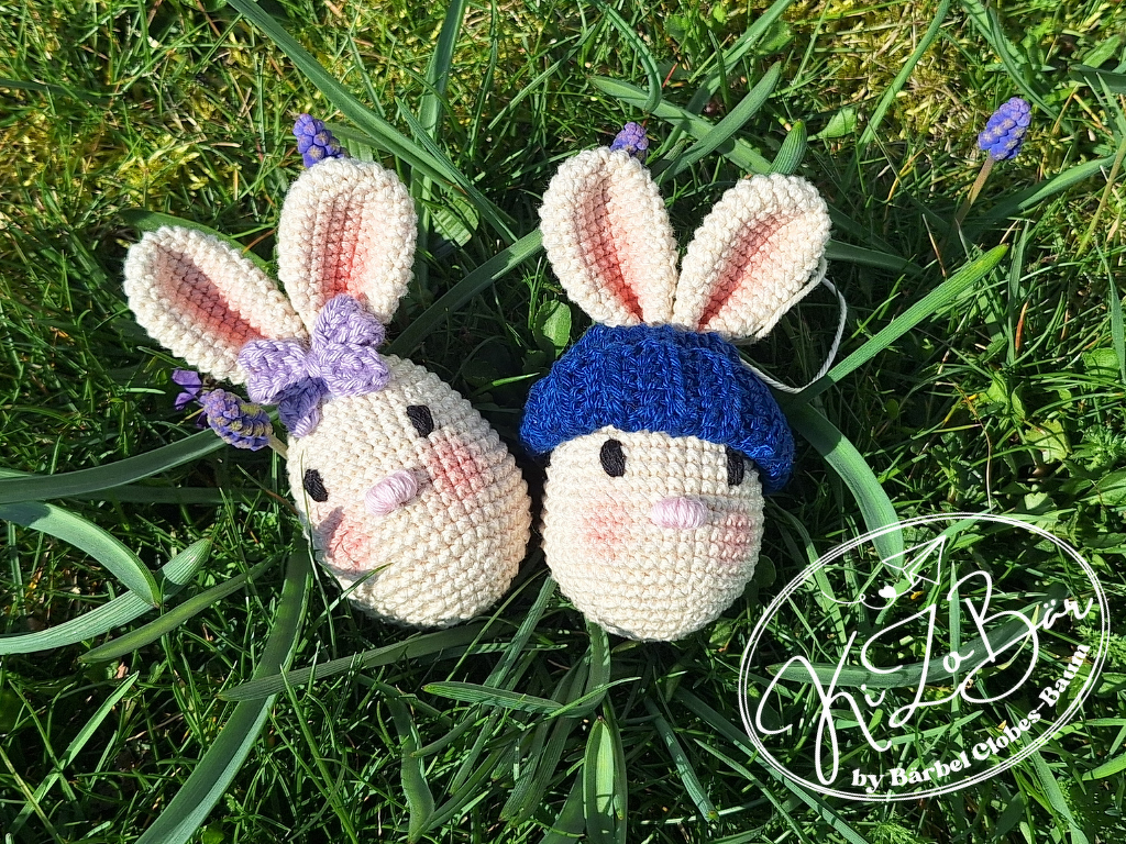 Zwei gehäkelte Hasen in Eierform im Gras, einer mit lila Schleife, einer mit blauer Mütze.