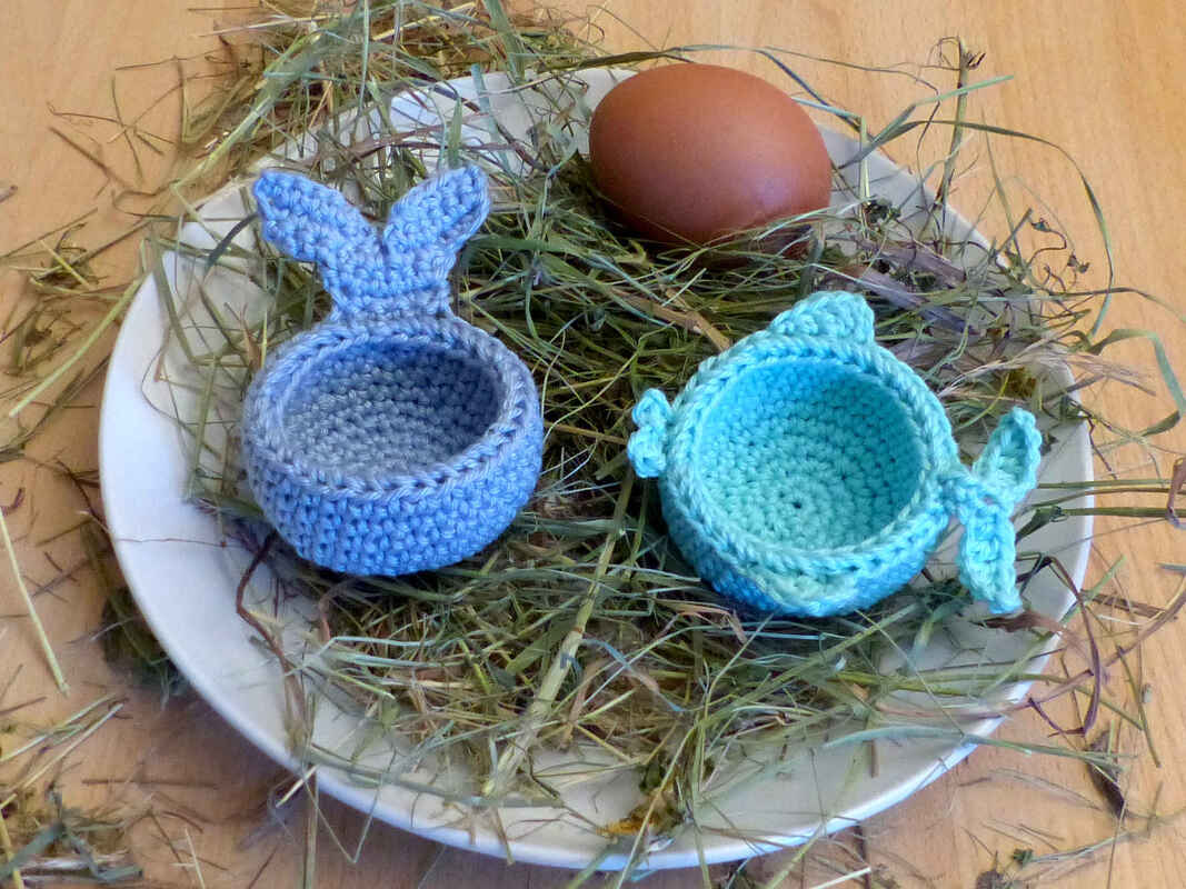 Zwei gehäkelte Eierwärmer in Blau: ein Hase und ein Wal, auf einem Teller mit Heu und einem braunen Ei