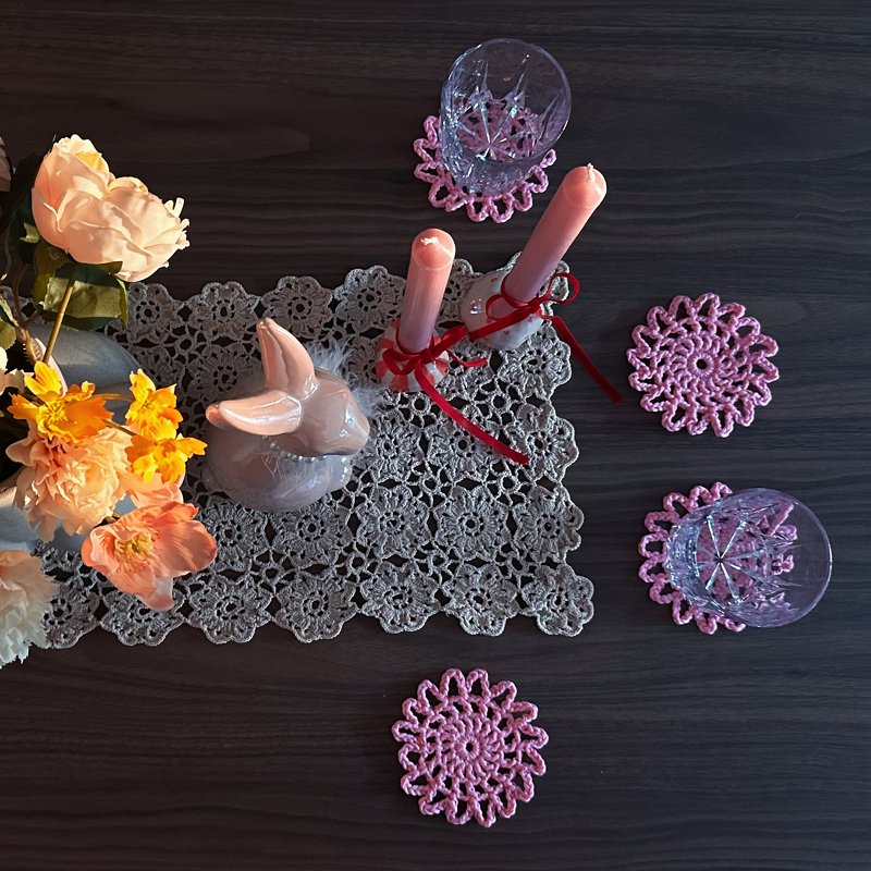 Vogelperspektive eines dunklen Holztisches mit grauem gehäkeltem Tischläufer, rosa Kerzen, rosa gehäkelten Untersetzern, Gläsern, Hasenfigur und Blumen.