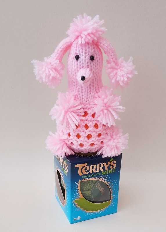 Pink knitted poodle cozy with yarn pompoms and black bead eyes on a blue box