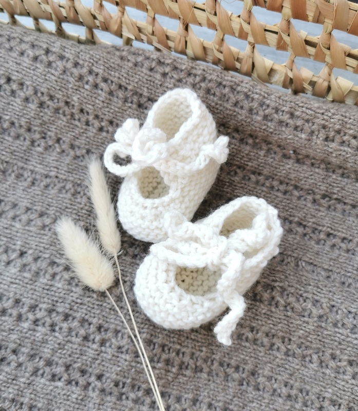 Weiße gestrickte Ballerina-Babyschuhe mit Bindeband auf brauner Strickdecke und getrockneten Gräsern
