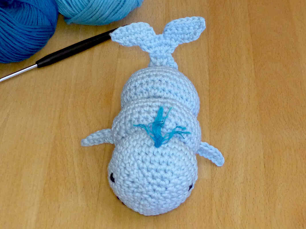 Hellblaues gehäkeltes Wal-Amigurumi neben Häkelnadel und Garn auf hellem Holztisch