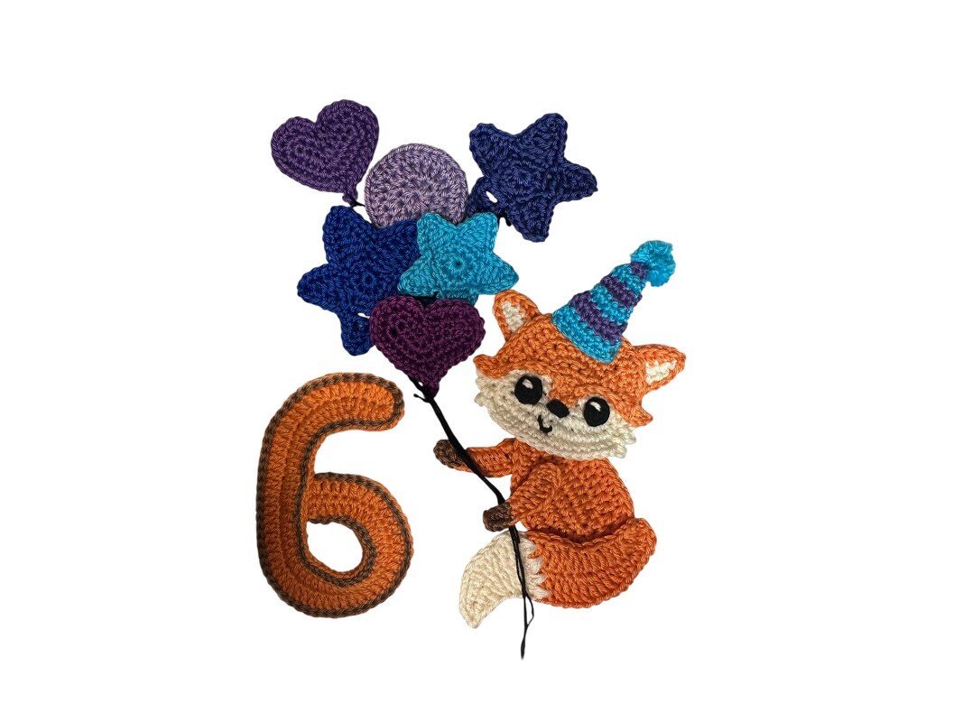 Crocheted orange fox wearing a blue-and-purple striped party hat, holding purple and blue heart and star balloons beside a crocheted number 6.
