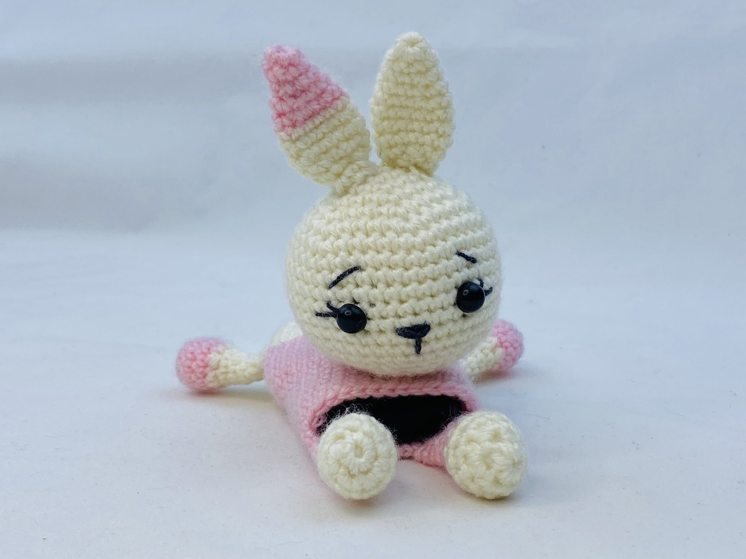 Cream crocheted amigurumi bunny with pink accents wearing a pink dress, sitting on a white surface.