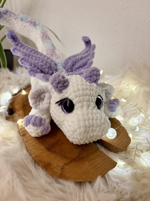 Gehäkelter weißer Drache mit fliederfarbenen Flügeln und großen dunklen Augen auf Holz- und Fellunterlage mit Lichterketten