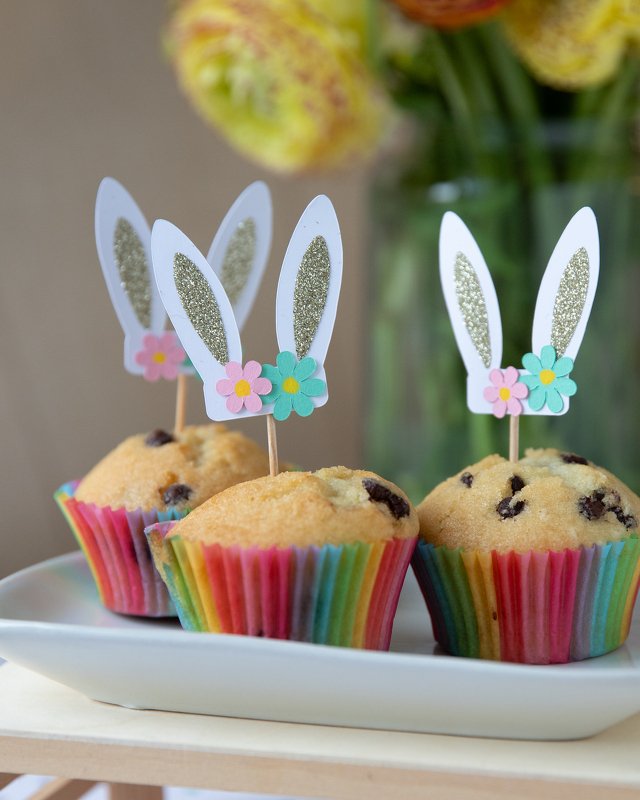 Detaillierte Ansicht von drei Cupcakes mit Papier-Hasenohren-Toppern, goldfarbenem Glitzer und kleinen Blumen.